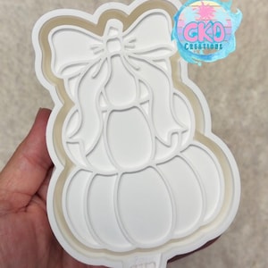 May include: A white plastic cookie cutter shaped like a stacked pumpkin with a bow on top. The design features a large bottom pumpkin, a smaller middle pumpkin, and a decorative bow at the peak. A circular logo with the text GKO CREATIONS is visible.