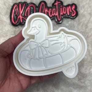 May include: White plastic duck-shaped cookie cutter with a bow and inner tube design. The cutter has a detailed outline of a duck with a bow tie, sitting in an inner tube. The text "CKD creations" is visible in the background.