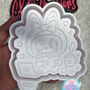 May include: A white, detailed mold featuring a cartoon character with a bow, working on a laptop and holding a phone. The mold has a scalloped edge with hearts, stars, and a diamond shape. The text "CKD" is visible on the mold.