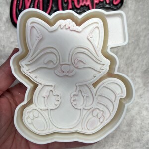 May include: White plastic cookie cutter shaped like a cartoon raccoon with a pink outline. The raccoon is sitting with its paws in front of it and has a striped tail.