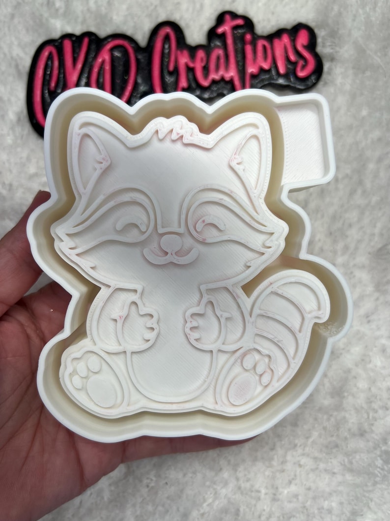 May include: White plastic cookie cutter shaped like a cartoon raccoon with a pink outline. The raccoon is sitting with its paws in front of it and has a striped tail.