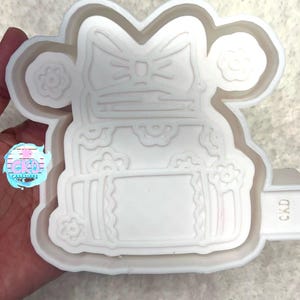 May include: White silicone mold of a cartoon character with a bow, holding a gift box with a bow. The mold is marked "CKD".