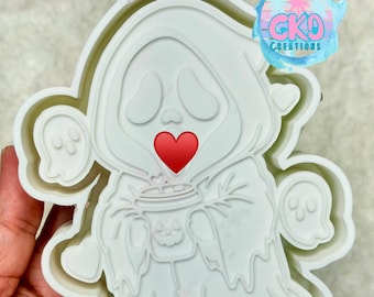 May include: A white plastic cookie cutter featuring a cartoon ghost character wearing a hooded cloak, holding a cup with a jack-o'-lantern face. The design includes two smaller ghosts and hearts on the sides. The text CKD is embossed at the top.