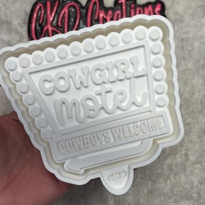 May include: A white plastic cookie cutter in the shape of a retro motel sign. The sign reads "Cowgirl Motel" with "Cowboys Welcome" below. The top of the sign has a series of small circles. The logo "CKD Creations" is visible in the background.