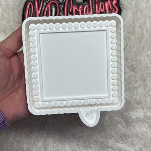 May include: A white square cookie cutter with a scalloped edge and a dotted border. The cutter has a rectangular center and a small tab on the bottom. The text "CKD" is visible on the tab. The logo "OVO Creations" is visible in the background.