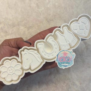 May include: A white plastic multi-shape cookie cutter featuring a daisy with a smiley face, a horse head, a horseshoe, a fringed vest, and a heart with a cow print pattern. A circular sticker with the text GKD CREATIONS is attached to the side.