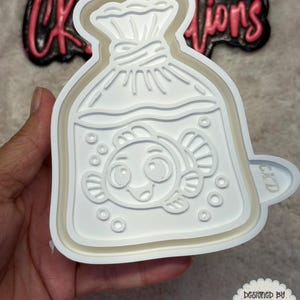 May include: A white plastic cookie cutter in the shape of a fish in a bag. The fish has a smiling face and is surrounded by bubbles. The cutter is held in a hand. The text "CK Creations" is in the background.