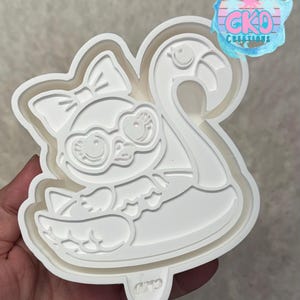 May include: A white plastic cookie cutter in the shape of a cartoon flamingo wearing heart-shaped glasses and a bow. The flamingo is floating on an inner tube. The cutter has the text "CKD Creations" on the top right.