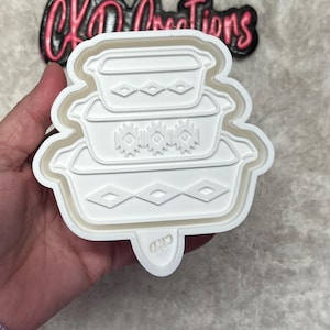 May include: A white plastic cookie cutter in the shape of three stacked casserole dishes with diamond and floral designs. The cutter is held in a hand. The text "CKD Creations" is in the background.