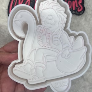 May include: A white plastic cookie cutter in the shape of a person riding a swan. The person is wearing glasses and a button-up shirt. The cookie cutter has a decorative outer edge and the letters "CKD" are visible.