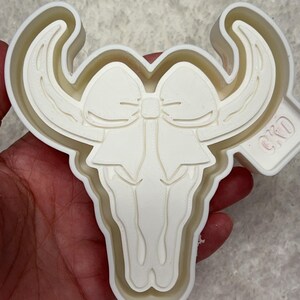 May include: White plastic cookie cutter shaped like a bull skull with a bow on top. The cutter has a raised edge and the text "CRD" is visible on the side.