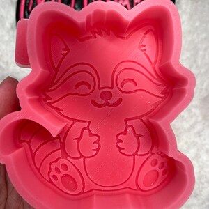 May include: Pink silicone mold of a cartoon raccoon with a happy expression. The raccoon is sitting with its paws in front of it. The mold is designed for making resin crafts.