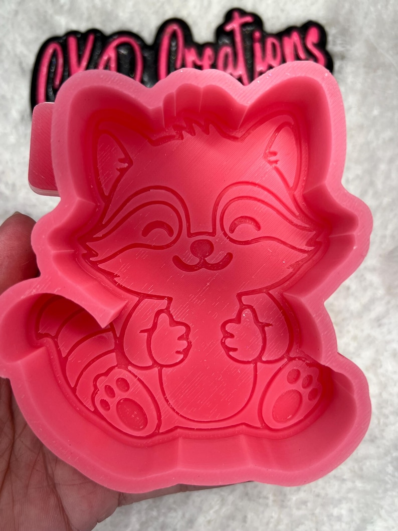 May include: Pink silicone mold of a cartoon raccoon with a happy expression. The raccoon is sitting with its paws in front of it. The mold is designed for making resin crafts.