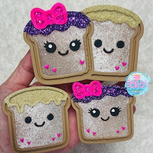 May include: Four brown bread-shaped charms with glittery details. Two charms have pink bows and purple glitter, while the other two have gold glitter. Each charm has a smiling face with hearts.