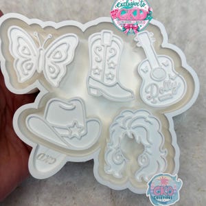 May include: A white silicone mold featuring five distinct cavities: a butterfly, a cowboy boot with star details, a guitar with the word Dolly, a cowboy hat with a star, and a voluminous hairstyle. The mold includes two CKD Creations logos.