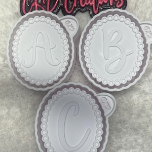 May include: Three white letter-shaped cookie cutters with a scalloped edge and a decorative border. The letters A, B, and C are visible. The text "CKD Creations" is above the cookie cutters.