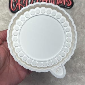 May include: A white, circular cookie cutter with a scalloped edge and a dotted border. The center is a smooth, blank circle. A small tab extends from the side. The text "CKD Creations" is visible in the background.