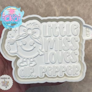 May include: White plastic mold featuring the words "Little Miss Loves Dr. Pepper" with an illustration of a smiling character holding a mug. The mold has a layered design and is likely for crafting or baking.