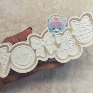 May include: A white multi-shape cookie cutter featuring a checkered lightning bolt, a smiley face with lightning bolt eyes, a bow, a pair of cherries with a bow, and a disco ball with a bow. The cutter includes a logo sticker reading CKD CREATIONS.