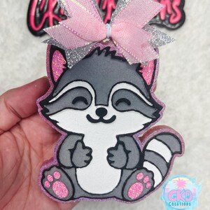 May include: A gray raccoon with a pink bow on its head. The raccoon is sitting with its paws in front of it. The raccoon is made of foam and has glitter on it.