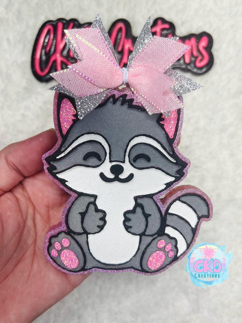 May include: A gray raccoon with a pink bow on its head. The raccoon is sitting with its paws in front of it. The raccoon is made of foam and has glitter on it.