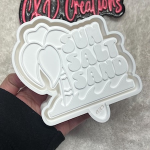 May include: White plastic cookie cutter with the words "SUN SALT SAND" in a retro font, along with a palm tree and waves. The cutter is held in a hand. The logo "CKD Creations" is visible.