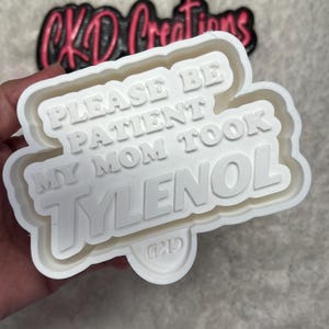May include: A white plastic cookie cutter with the text "PLEASE BE PATIENT MY MOM TOOK TYLENOL" in bold, capital letters. The cutter has a rounded rectangular shape with a small, circular detail at the bottom. The background features a blurred logo.