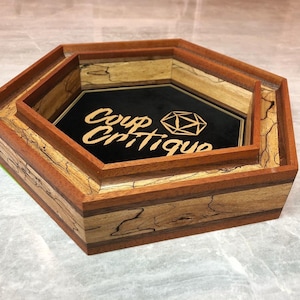 May include: A hexagonal wooden dice tray with a black felt bottom and a wooden inner tray. The tray has a natural wood finish with brown and tan stripes. The tray is engraved with the text "Coup Critique" and a stylized dice icon.