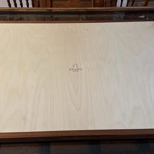 May include: A rectangular wooden frame with a light-coloured wood panel. The frame is dark brown with brass-coloured corner hardware. The panel has a wood grain pattern and a small logo that reads "BLACK BEAR".
