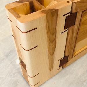 May include: A wooden knife block with a folding design. The block has multiple slots for knives and a separate compartment for other utensils. The block is made of light wood with dark wood accents.