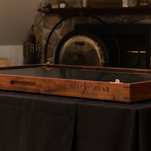 May include: A rectangular wooden display case with a glass top and metal latches. The side of the dark brown wood frame features the engraved text Black Bear Workshops. The case has integrated side handles and a smooth, polished finish.