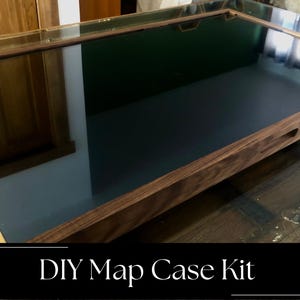 Pode incluir: Uma caixa de exposição retangular de madeira com topo de vidro e base interior escura. A moldura apresenta um acabamento de grão de madeira natural e uma pega recortada na lateral. O texto DIY Map Case Kit está em branco numa faixa preta.