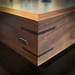May include: Close-up of a wooden box with dovetail joints and a metal accent. The box is made of dark brown wood with lighter wood trim. A small, oval-shaped metal piece is visible on the side of the box.