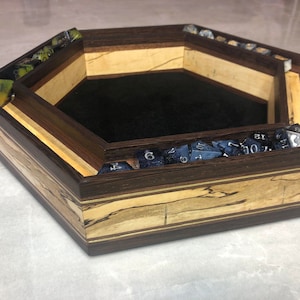 May include: A hexagonal wooden dice tray with a black felt interior and a separate compartment for dice. The tray is made of dark wood with light wood accents and has a natural wood grain pattern.