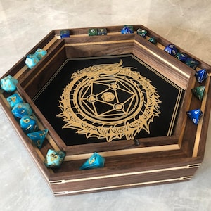 May include: A wooden hexagonal dice tray with a black felt bottom featuring a gold dragon design. The tray has compartments around the edge for dice and a central area for rolling. The tray is filled with blue and green dice.