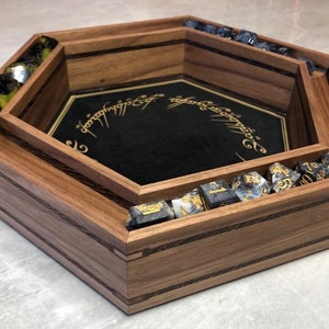May include: A hexagonal wooden dice tray with a black felt bottom featuring the inscription "One Ring to rule them all". The tray has a raised inner rim and is made of walnut wood with a striped pattern.