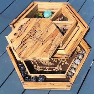 May include: A hexagonal wooden dice tray with a removable lid. The lid features a woodgrain pattern and the text "Black Bear Workshops" burned into the wood. The tray has a brown felt lining with a gold tree design. The tray is filled with black and blue dice.