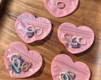 Peut inclure: Six pochettes zippées en forme de cœur rose avec des fenêtres en plastique transparent, chacune contenant une variété de bracelets en perles colorées.