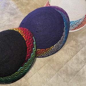 May include: Three crocheted kippahs with colourful geometric patterns. The kippahs are navy blue, black and white.