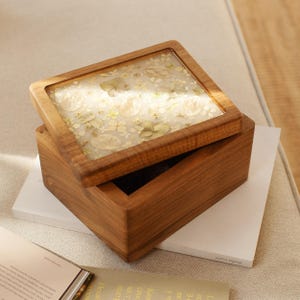 Peut inclure: Une boîte en bois avec un couvercle à charnière, avec un dessus transparent incrusté de fleurs séchées. La boîte est ouverte, révélant l'intérieur. Le bois a une teinte chaude et naturelle. La boîte est posée sur une surface blanche.