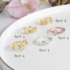May include: Five gold rings with different names engraved on them in different fonts. The names are "Sarah", "Stephanie", "Betty", "Emma", and "Nicole". The rings are displayed on a white plate with the font number written next to each ring.