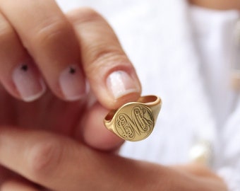 May include: A gold signet ring with an oval face featuring an engraved monogram. The ring is held up against a white background. The ring's design is detailed and elegant, showcasing a classic jewelry style.