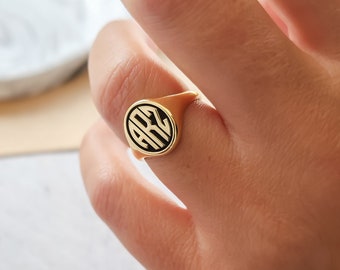 May include: A gold-colored signet ring with a black circular design featuring the letters "ARZ". The ring has a smooth, polished finish and is worn on a finger. The ring's design is centered on the face of the ring.