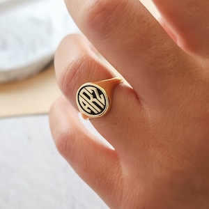May include: A gold-colored signet ring with a black circular design featuring the letters "ARZ". The ring has a smooth, polished finish and is worn on a finger. The ring's design is centered on the face of the ring.
