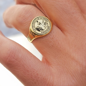 May include: A gold ring with a black and white seal featuring the University of Alabama at Birmingham logo. The ring has the initials "ANK" engraved on the band.