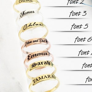 Può includere: Sette anelli d'oro con diversi nomi incisi, tra cui Olivia, Shomas, Chloc, Mom and Dad, Emma, Sarah e TAMARA. Gli anelli sono disposti verticalmente, con i nomi rivolti verso lo spettatore. Gli anelli sono su uno sfondo bianco con una pianta blu e verde sullo sfondo.