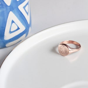 May include: A rose gold signet ring with a monogrammed oval top. The ring is sitting on a white plate.