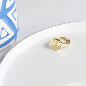 May include: A gold signet ring with a cursive letter "F" engraved on the top. The ring is on a white plate.