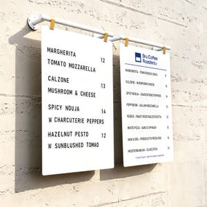 May include: Two white rectangular menu boards suspended from a white horizontal bar with gold clips. The boards feature black text listing food items like Margherita, Calzone, Spicy Nduja, and Hazelnut Pesto, along with prices and Bru Coffee Roasters branding.