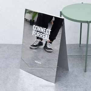 Puede incluir: Un letrero en forma de A con espejo con el texto "CONNECT FORREAL" en blanco. El letrero refleja a una persona que lleva pantalones negros y zapatillas negras y blancas. El letrero también tiene el texto "U.B. APP" en la parte inferior.